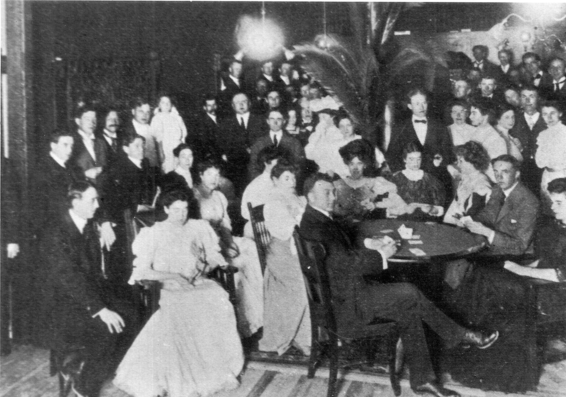 A large crowd of men in evening dress and women in long gowns packed into a hall around tables